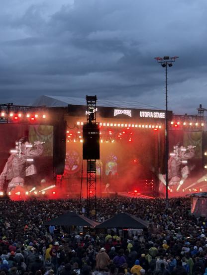 A large crowd gathers at a music festival under cloudy skies. The stage, labeled "UTOPIA STAGE," is lit with red lights and displays visuals on a backdrop, while performers are visible on stage, engaging the audience.