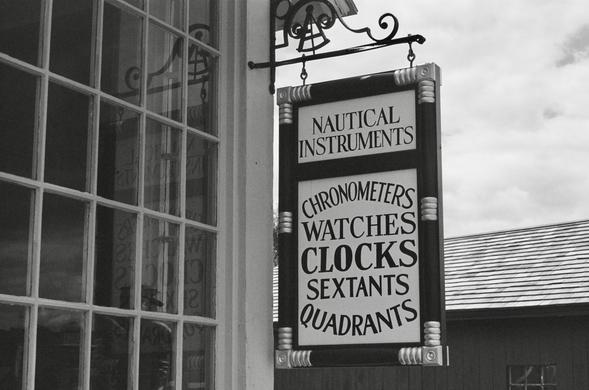 Black and white photo of a hand crafted wooden sign with black trim, carved gold painted trim, and old style caps black lettering on a white background. The top section of the sign reads: Nautical Instruments. The bottom section reads: Chronometers, Watches, Clocks, Sextants, Quadrants. The words Chronometers and Quadrant arc above and below the other words in the bottom section. The sign hangs from a wooden paned display window of a reconstructed instrument shop at Mystic Seaport Museum, Mystic CT.