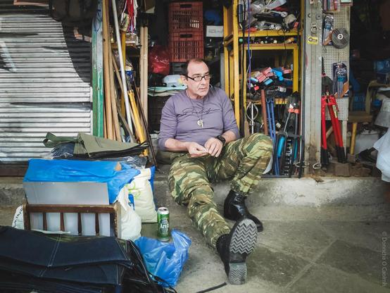 A candid street photo taken in Athens, Greece, showing a man in camouflage trousers and black boots sitting on the ground outside a cluttered tool shop, surrounded by bags, a can of Areti beer, and various hardware items, with shelves full of tools and equipment in the background.