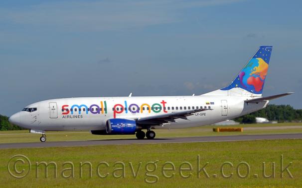 Side view of a twin engined jet airliner accelerating from right to left along a black runway.
The plane is mostly white, with multi-coloured "Small Planet" titles along the fuselage, successive letters in red, blue, purple, yellow, and green.
The black registration "LY-SPE" is on the lower rear fuselage.
The engine pods mounted under the wings are a deep blue, with white "SmallPlanet.Aero" text.
The tail is dark blue, with a stylised multi-coloured globe and yellow stars.
Lush green grass fills the foreground, with tall green trees in the distance behind the plane on either side, under blue sky with smears of cloud.