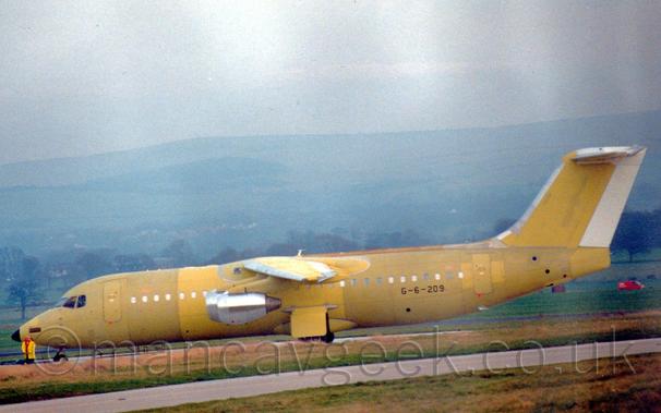 Side view of a high-winged, 4 engined jet airliner parked facing to the left.
The plane is mostly yellow, with various barrel sections of the fuselage and wings and different panels painted in different shades.
The engine pods are bare metal, and the rudder on the tail is plain white.
The black registration "G-6-209" is on the lower rear fuselage.
Green grass lines a taxiway in the foreground, with more grass leading up to trees marking the edge of the airfield in the background beyond that, and rolling hills in the distance, under grey skies.