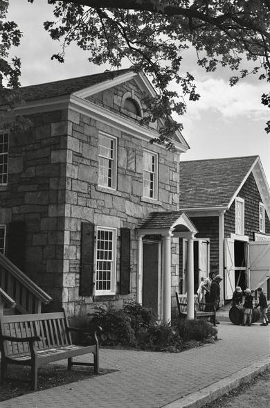 Black and white photo of Mystic Bank, 1856. A two-story cut stone commercial bank built in the Greek Revival style. The front of the bank has two white-framed multipane windows on the second floor. The first floor has a white-framed multipane window with black shutters on the left, next to a door within a small open portico supported by two Doric columns. The white-framed pediment of the roof has a small half-round cut stone window in centered in the pediment. Ship builders and buyers would trust this bank! Moved from Old Mystic to Mystic Seaport Museum, Mystic, CT in 1951.
