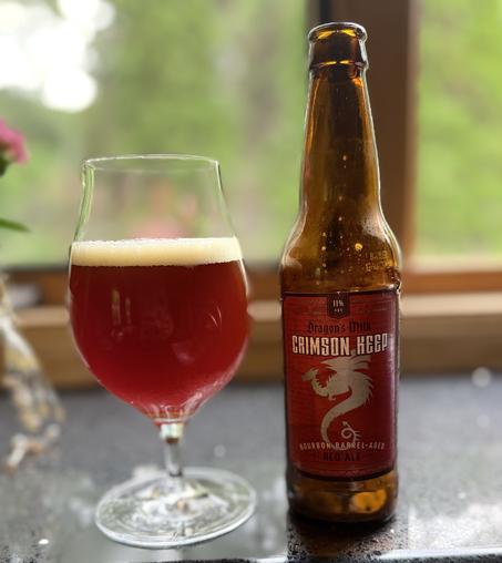 A snifter of ruby red beer next to a brown bottle with a dark red label and a silver dragon in silhouette