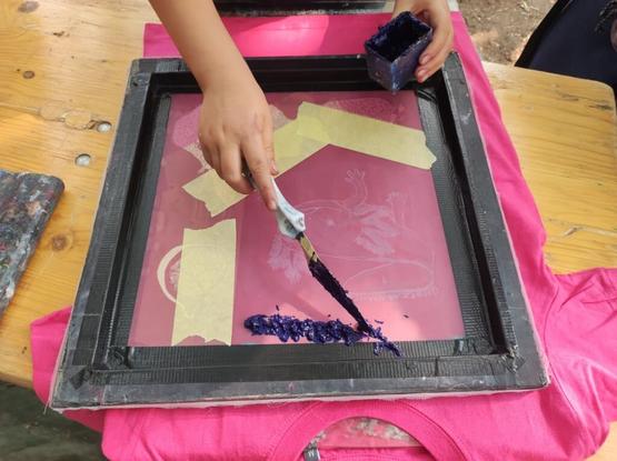 A photo of hands spreading dark blue color on a screen, underneath the screen is a pink shirt