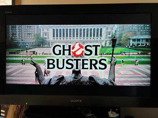 A TV showing the opening titles of the movie Ghostbusters. The movie logo is at the centre (on two lines weirdly as everywhere else has referred to it as one word) on top of an outdoor shot in New York City, Columbia University I believe.