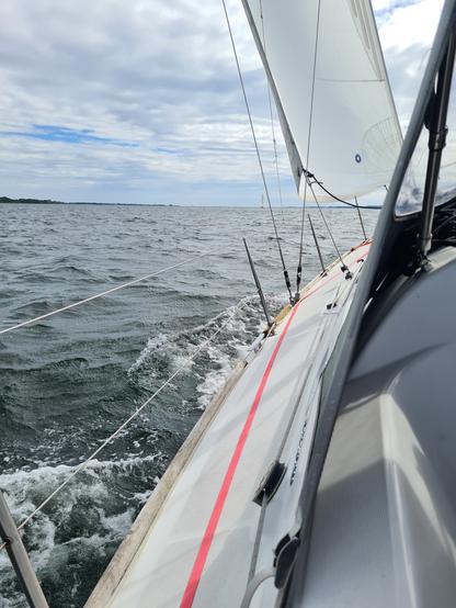 Blick über die Reling einer Segelyacht nach vorn, das Wasser schäumt am Rand des Boots, am Horizont ist Land zu sehen