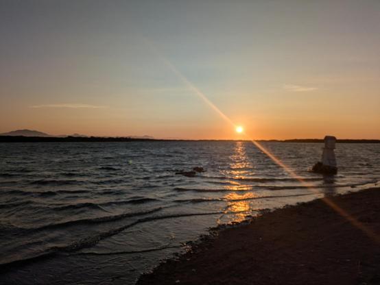 Atardecer en la costa pacifica mexicana. Se ven olas pequeñas, una isla al fondo y el sol ya muy cerca del horizonte. Del lado derecho de la foto se ve un pequeño altar en el mar.
