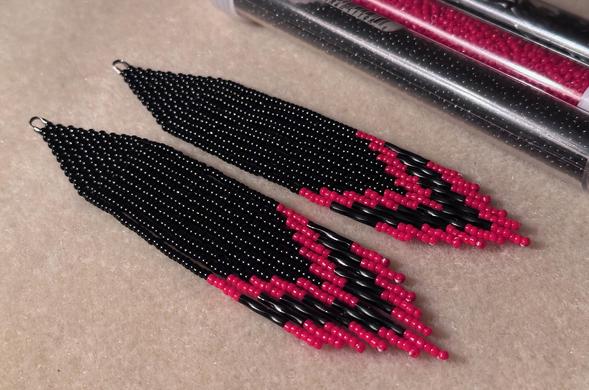 A pair of black beaded fringe earrings with red chevron stripes at the bottom
They are laying on a bead mat next to tubes of red and black seed beads