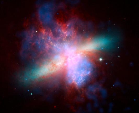 Composite of images of the active galaxy Messier 82. In the center of the image is a long cigar-shaped green and white light. In the center going up and down the image are clouds of gas/dust in red and blue.