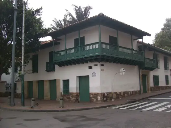 The image depicts a two-story building with a traditional architectural style, characterized by white walls and green wooden accents, including balconies and doors. The building appears to be located on a street with trees and includes some street signage and a crosswalk. The surroundings suggest a historical or cultural area, likely in a town or city that values its heritage.

Image Credits: Wikimedia / Baiji / CC BY-SA 4.0
