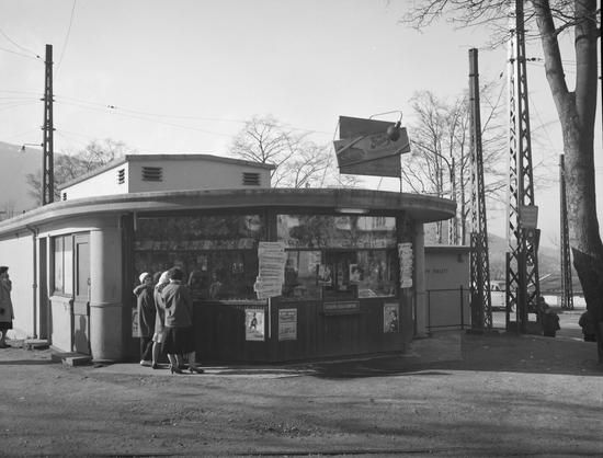 A black and white photo of a newspaper and magazine kiosk, some people gathered outside, advertisements for gossip magazines and chocolates.