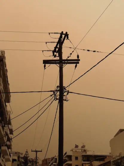 An utility pole in front of a desert yellow sky and houses to the left and bottom.