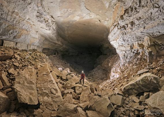 A person in a red cave oversuit and helmet with light on stands on a boulder just below middle of the photo. The ceiling is clearly at least 30 ft / 10m above them, if not more, and gets larger beyond where they stand as it fades into darkness. The ceiling is mostly smooth with patches of different coloration of limestone, some more gray and some more reddish. The walls show various layers, again some more red and some more gray. The floor is simply boulders and rock, ranging from basketball to a small vehicle in size.