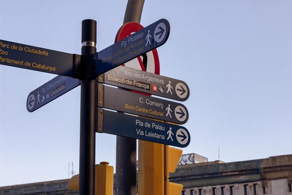 A pole with several wayfinding signs pointing towards different places of interest and directions. On one of these signs, the old Renfe logo can be seen, next to the name "Estació de França". Behind these signs, there's a traffic light.