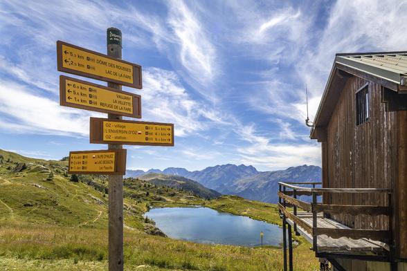 View from L'Alpette (2000m altitude) in Alpes d'Huez area with wooden signs showing hiking paths directions and a little wooden house in the foreground and a small turquoise blue mountain lake and mountain peaks in the background under a very blue sky with stretched white clouds.