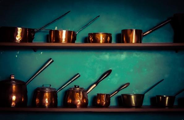 Copper cookware, including pots and pans, are neatly arranged on two stacked shelves against a deep turquoise wall. Each piece has a handle and varies in size and shape, reflecting ambient light with a warm glow.