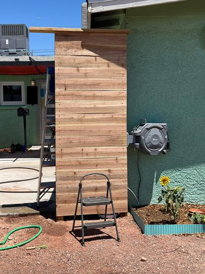 Tall and narrow wooden fence with a retractable hose attached in front of a green house.