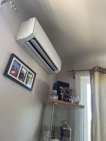 A register for the heat pump is installed on the top of a wall. There is a small picture frame below it and a glass shelf in the corner beside the window.