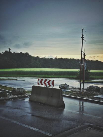 The portrait-orientated image shows an outdoor scene featuring a wet asphalt surface, with visible reflections indicating recent rain. In the foreground, a concrete barrier with a red and white chevron arrow sign pointing to the left is centrally placed on the asphalt. To the left and right of the barrier, several large, light-colored rocks are visible on the ground.
In the midground, a wide expanse of green, possibly a field of crops or grass, stretches horizontally across the frame. Beyond the green field, a dense line of dark green trees forms a horizon. The sky above is overcast with varying shades of gray, though a lighter, yellowish hue is visible towards the right side, suggesting the setting sun. On the right side of the image, a tall, dark pole with some attached equipment stands vertically. The overall lighting suggests dusk of an overcast day.