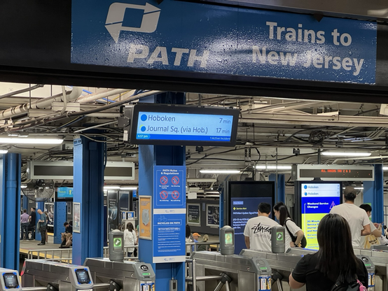 A blue and white lettered sign reading “PATH Trains to New Jersey” with some people entering the turnstiles.