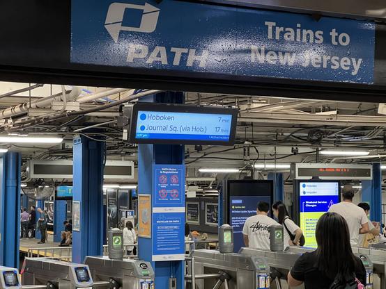 A blue and white lettered sign reading “PATH Trains to New Jersey” with some people entering the turnstiles.