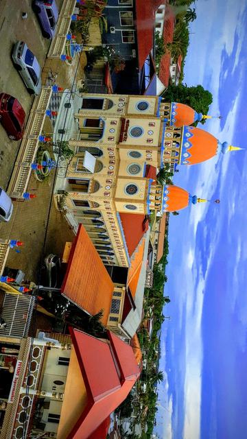 The photograph depicts a vibrant scene with a prominent building featuring bright orange domes and blue accents, suggesting a Mandir Mosque or a similar structure. The building is adorned with intricate patterns and has multiple arched windows. In the foreground, there are several parked cars, including a purple van and a red car, along with a red-roofed building labeled "FUHUANG Supermarket." The street is lined with a mix of residential and commercial buildings, some with red roofs, and a decorative fence with blue and red elements. The sky is clear with a few scattered clouds, and the surrounding area is lush with greenery, including palm trees. The overall atmosphere is lively and colorful, with a blend of architectural styles.

 Ovis2-8B

🌱 Energy used: 0.185 Wh
