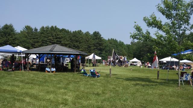 People with open tents at a PowWow. The grass is green, the sky is hazy and sort of blue. There are pine trees in the distance.