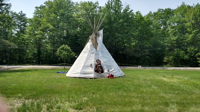 A teepee near the entrance of a PowWow. The grass is green, the sky is hazy and sort of blue. There are pine and deciduous trees in the distance.