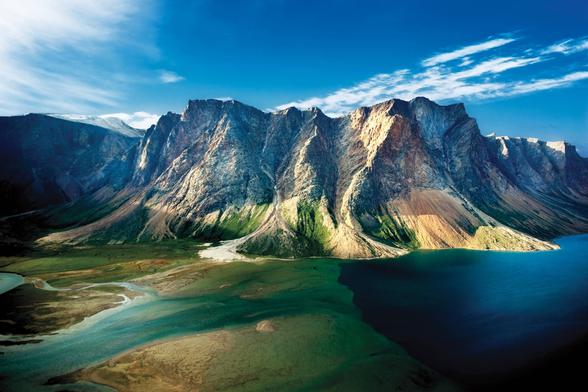 An aerial photograph showcases the rugged beauty of the Torngat Mountains in Labrador. Towering, layered rock formations rise dramatically from a turquoise fjord under a clear blue sky.