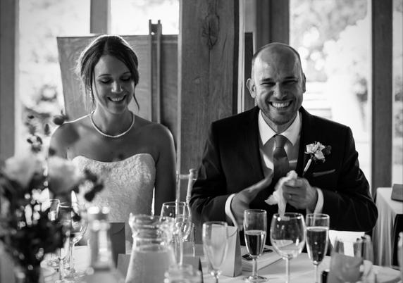 Bride and groom seated at a wedding reception table, both smiling. The groom wears a dark suit and tie with a boutonniere and holds a napkin in his hands. The bride wears a strapless lace dress and a pearl necklace. Several glasses and floral decorations are visible in the foreground.