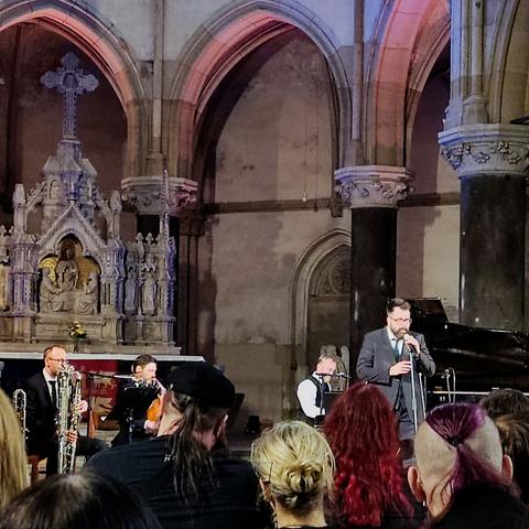 Blick in den Altarraum einer Kirche, darin 4 Personen: einer Sänger, ein Pianist, ein Violinist und ein Blechbläser. Davor eine Menge Personen, die aufmerksam zuhören