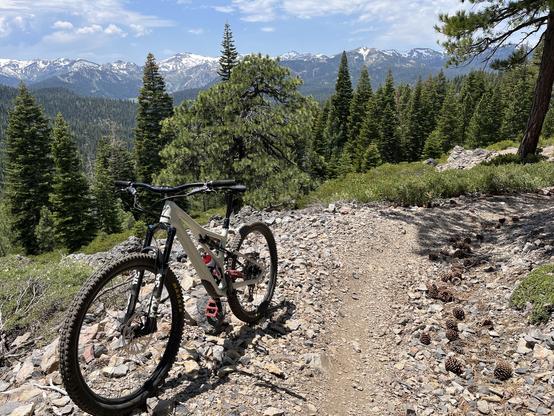 Mountain bike on side of a rocky trail with the snow-capped mountains of Palisades Tahoe  in the background