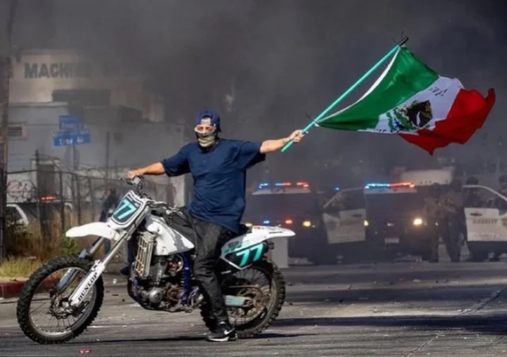Persona con googles y una gorra ondeando una bandera de mexico arriba de una motocicleta en frente de ICE