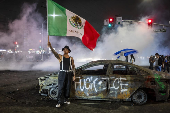 Persona con atuendo cholo clásico con sombrero y una bandera de México en frente de un carro quemado que dice: Fuck ICE