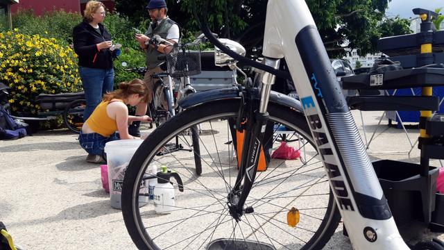 Une jeune femme entretien son vélo qui a passé tout l'hiver dehors tandis qu'une femme et un homme causent patins de frein à l'arrière plan. Un VAE attend son tour au 1er plan ;-)