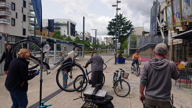 Atelier Hérouelibre en pleine effervescence devant le Café des Images. À gauche une femme nettoie son plateau avant, au centre un homme observe un vélo, et au loin une jeune femme teste son dérailleur fraîchement réglé. Un homme de dos observe la scène.