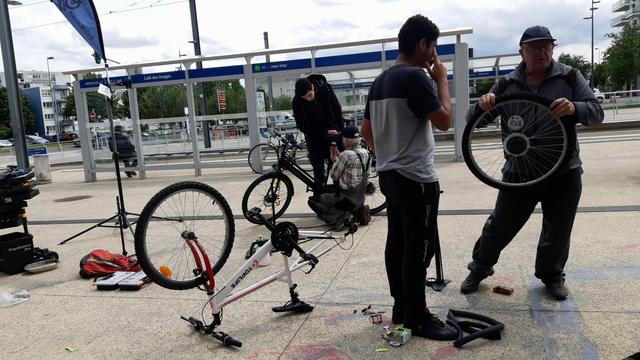 Au centre devant un arrêt de tram deux hommes travaillent sur un VAE, tandis que deux autres à droite tentent une danse avec une roue (et pas avec les loups) ;-)