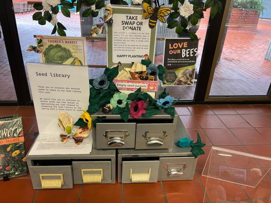 A display from the public library, featuring a 5-drawer card catalogue which has been repurposed as a seed library, plus paper flowers and butterfly. There are various notices. One reads:
Seed Library
Thank you to everyone who donated seeds to share in our Seed Library this year.
We would love you to continue to donate seeds now that Autumn is approaching, and look forward to reopening the Seed Library in the Spring with a fresh batch of seeds.

Another notice reads:
Take swap or donate
Help yourself to something from the Seed Library! You can take something away to plant, swap some of your leftover seeds for something else you need, or donate seeds you don’t need!
Make sure to put the seeds into an envelope and label the envelope with what the seeds you’re leaving will grow into!
Happy planting!