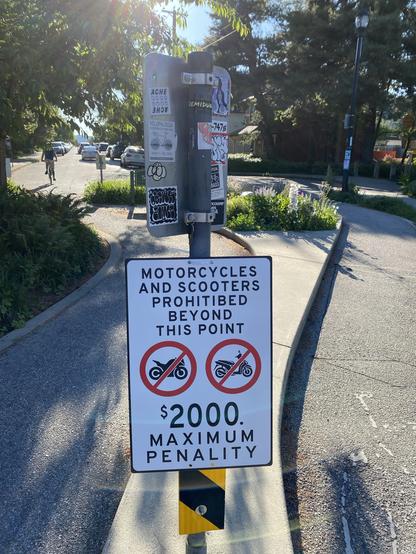 street sign in my neighbourhood, reading "MOTORCYCLES AND SCOOTERS PROHITIBED [sic] BEYOND THIS POINT", followed by red circled and crossed out images of a Ducati-type motorbike and a Yamaha-type scooter, followed by "$2000 MAXIMUM PENALITY [sic]"
