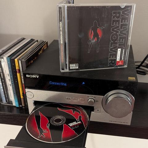 Un sistema de audio Sony CMT-SX7 reproduce un CD de Velvet Revolver, titulado "Contraband". La caratula del CD, que está en una bandeja de carga, y la caja del CD muestran una silueta de una mujer con una pistola. Detrás del sistema de audio hay una colección de otros CDs.