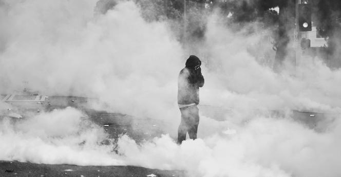 A hooded figure in black stands clutching their face in clouds of white smoke, presumably tear gas