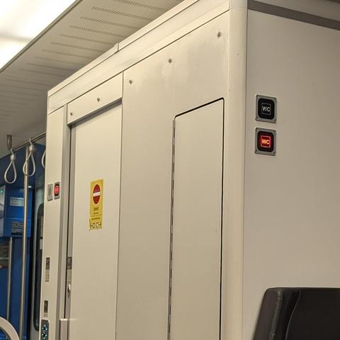 Eine weiße Zugtoilette mit rot leuchtendem „WC“-Symbol, das Belegung anzeigt. Ein gelbes Schild mit „Defekt“ und Verbotsschild ist an der Tür angebracht. Ein Teil eines Sitzes und Haltegruffe im Zug sind sichtbar.