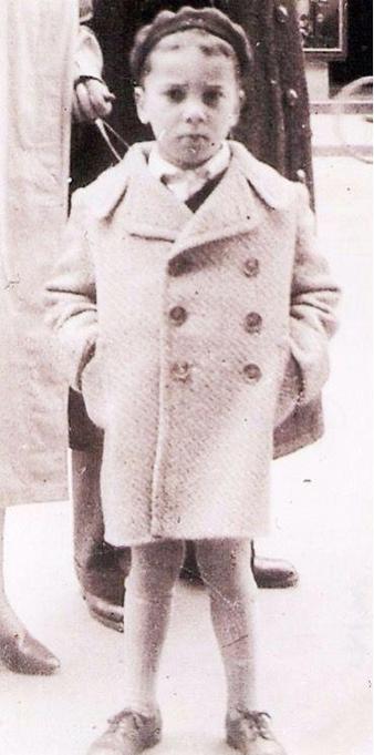 A young boy stands dressed in a double-breasted coat and shoes, expressing a serious demeanor, in a black and white historical photograph.