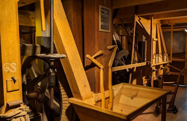 Blick in eine Getreide-Mühle. Im Vordergrund ein hölzerner Trichter, in den das angelieferte Korn geschüttet werden kann. Im Hintergrund gußeiserne Kammräder und Transmissionsriemen. Eine Sackkarre steht vor einer Absackanlage aus Holz.
