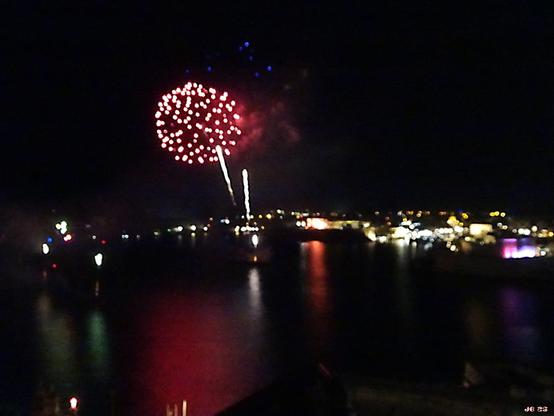 Nachtaufnahme von einem Feuerwerk in der Bucht von Valletta in Malta am 25.4.25. Zentrales Motiv ist eine Feuerwerkskugel, die aus vielen runden weißen Pünktchen zusammengesetzt ist. Unten das dunkle Meer, in der Mitte der beleuchtete Streifen von Gebäuden am anderen Ufer, oben der dunkle Himmel mit drei blauen Pünktchen vom vorausgehenden Feuerwerkskörper.