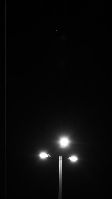 Streetlight with three illuminated lamps in the lower portion, against a dark night sky. A small bright star is visible above the streetlight, centered in a wide black expanse.