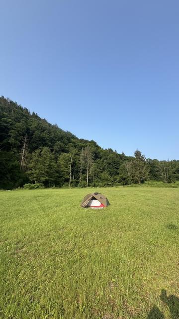 Ein kleines Zelt ist auf einem grünen Feld aufgebaut, umgeben von Bäumen und Hügeln unter einem klaren blauen Himmel.