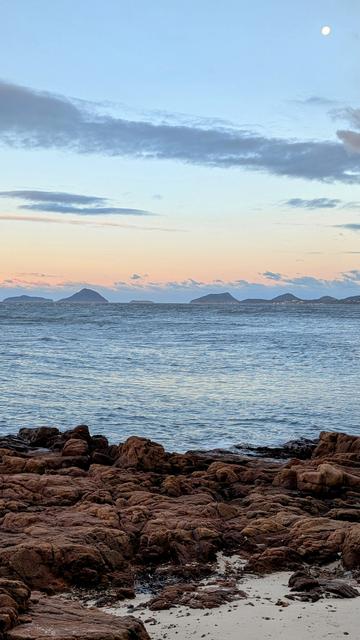 Looking across the waters of Port Stephens Bay to the entry to the ocean, which is flanked by twin small peaks on each side. An almost full moon is in a clear sky with a few whispy clouds. The water is a bit choppy due to strong westerlies. In the foreground the waters edge is flanked by fractured rocks.
