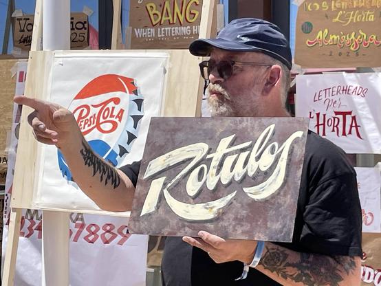 Man pointing at something out of the frame while holding a hand-painted signs that says "Rotulos".