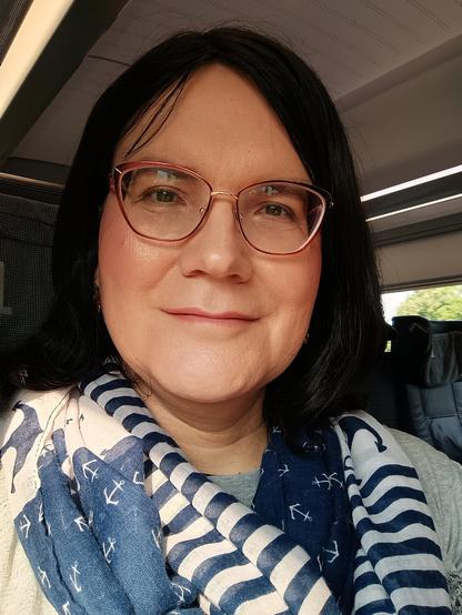 Ich, eine Frau mit dunklem schulterlangem Haar, Brille und blau-weiß gemustertem Halstuch mit Ankern, in einem ICE, lächelnd.
